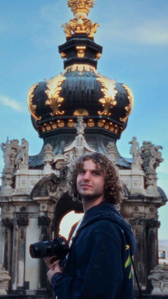 Pit Hantzsche mit einer Kamera in der Hand vor einem barocken Torbau mit vergoldeter Krone, dahinter blauer Himmel.