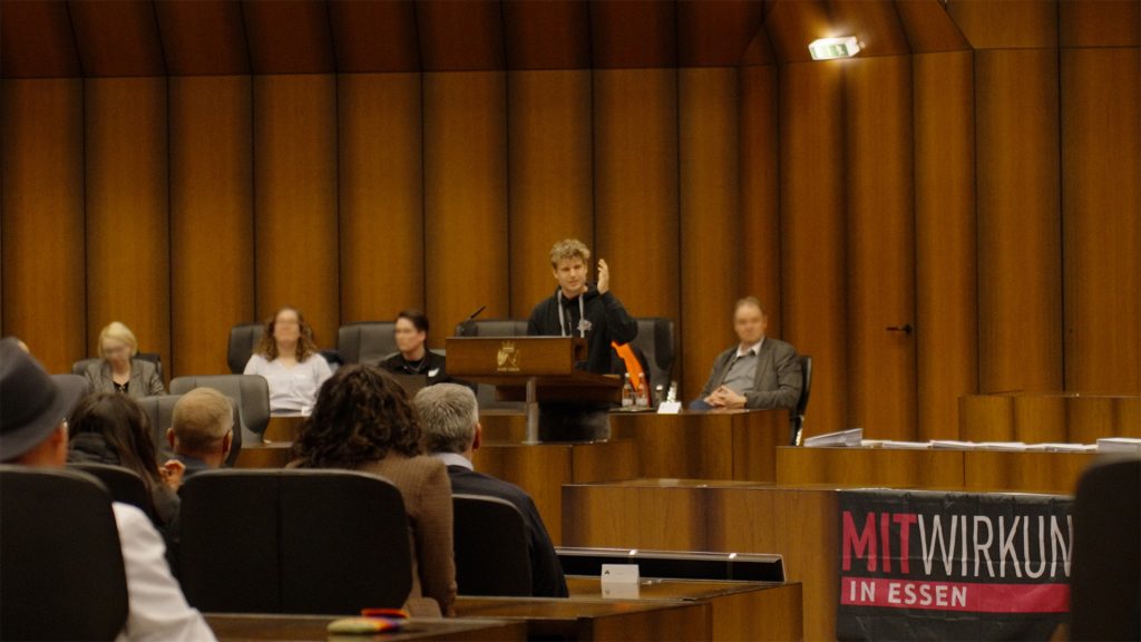 Rede am Podium im Ratssaal bei einer Veranstaltung von Mitwirkung in Essen, davor das Publikum und ein rotes Banner mit dem Schriftzug Mitwirkung in Essen.