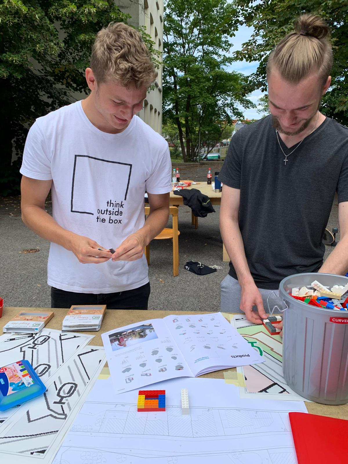 Zwei junge Männer stehen an einem Tisch mit Bastelmaterial und Plänen und arbeiten an einem Modell.