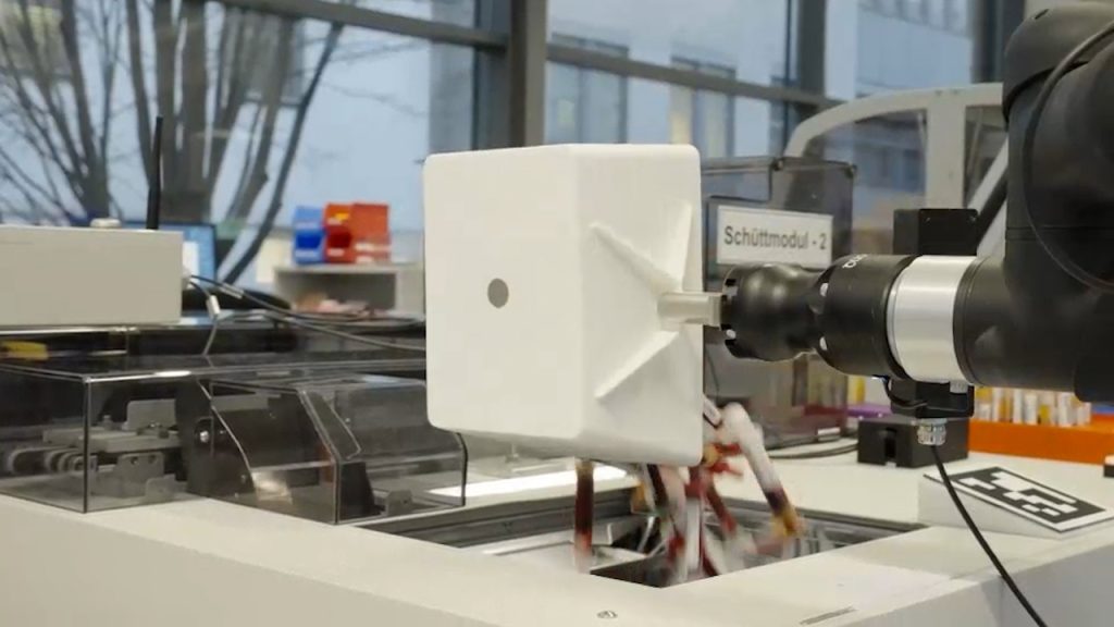 Roboterarm mit weißem Aufsatz über einer Laborbank im Zentrallabor, daneben Analysegeräte und Probenträger.