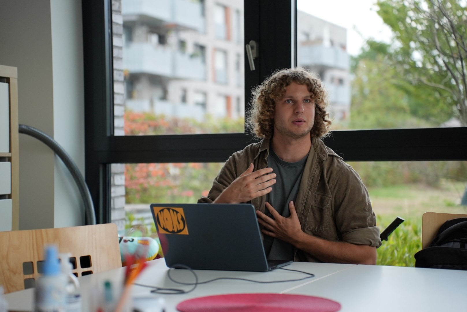 Pit Hantzsche sitzt an einem Tisch vor einem aufgeklappten Laptop und spricht mit erhobener Hand, im Hintergrund ein großes Fenster mit Blick auf Wohnhäuser.